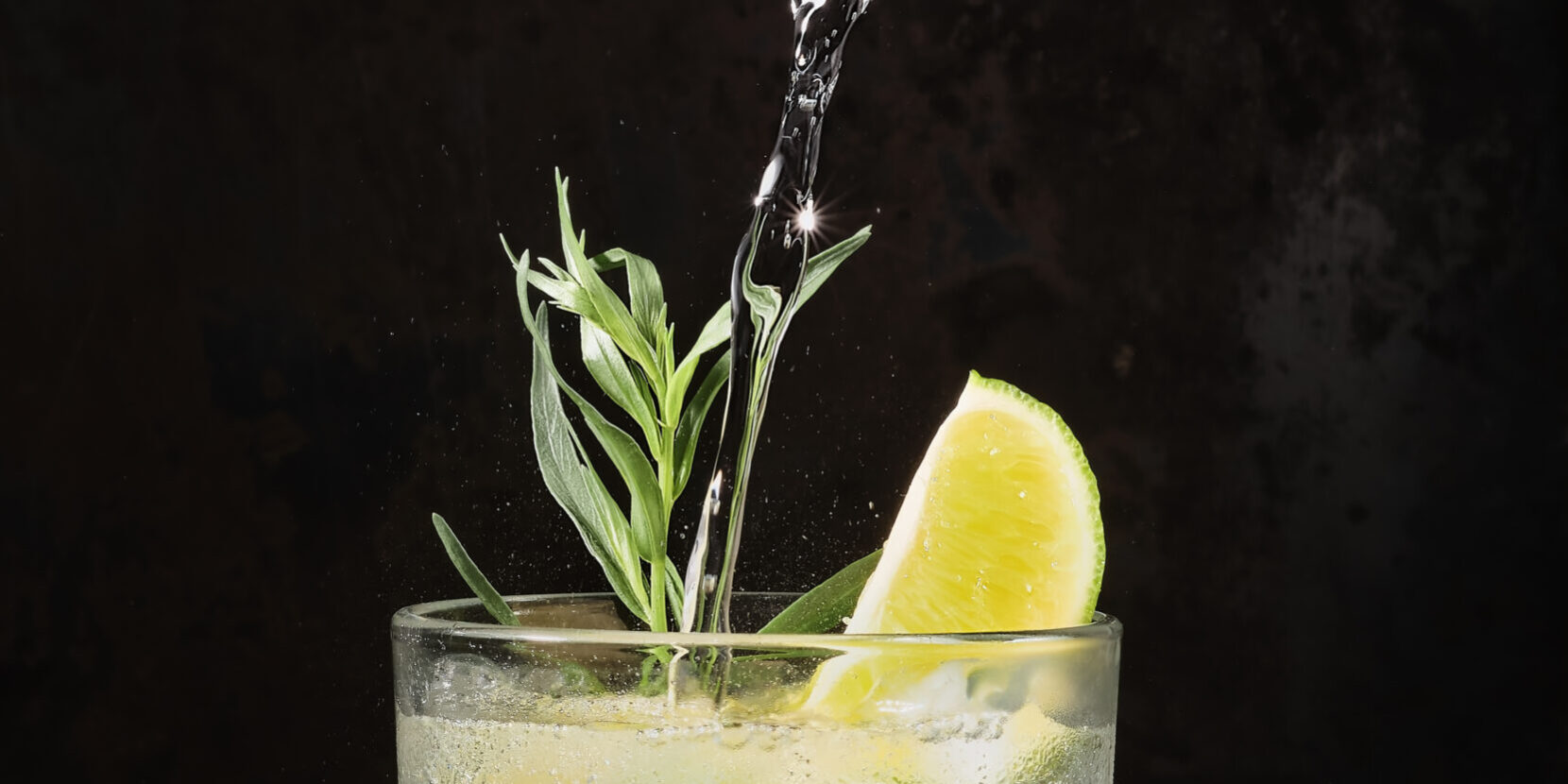 Glass of lime lemonade on dark table, summer drinks. Pure mineral water is poured into glass. Vertical frame, selective focus. Homemade drink with lime, tarragon and ice cubes. Cold Fresh Drinks idea Alcohol-free drinks