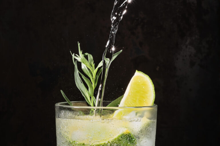 Glass of lime lemonade on dark table, summer drinks. Pure mineral water is poured into glass. Vertical frame, selective focus. Homemade drink with lime, tarragon and ice cubes. Cold Fresh Drinks idea Alcohol-free drinks